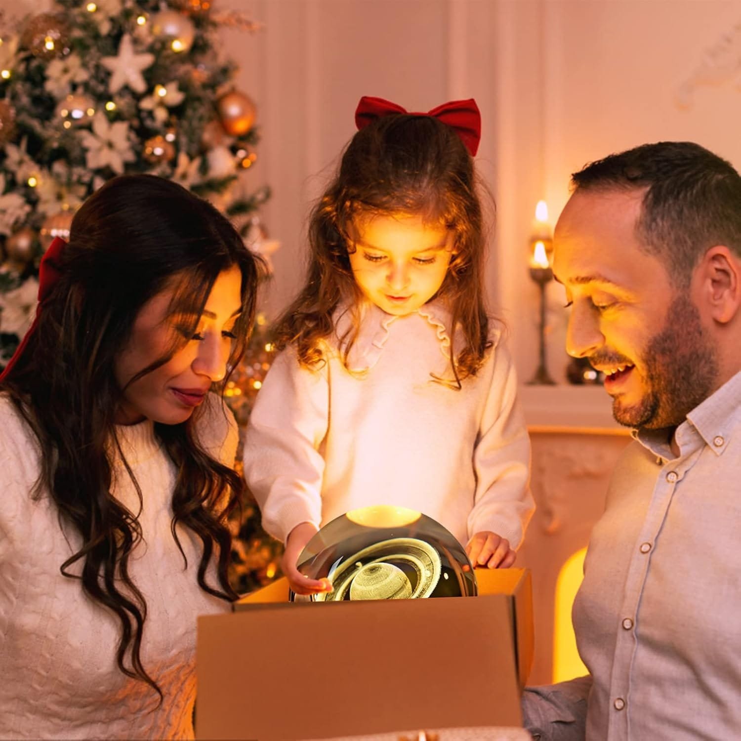 A family of three opens a glowing gift box in a festive room with a decorated Christmas tree and lit candles in the background.