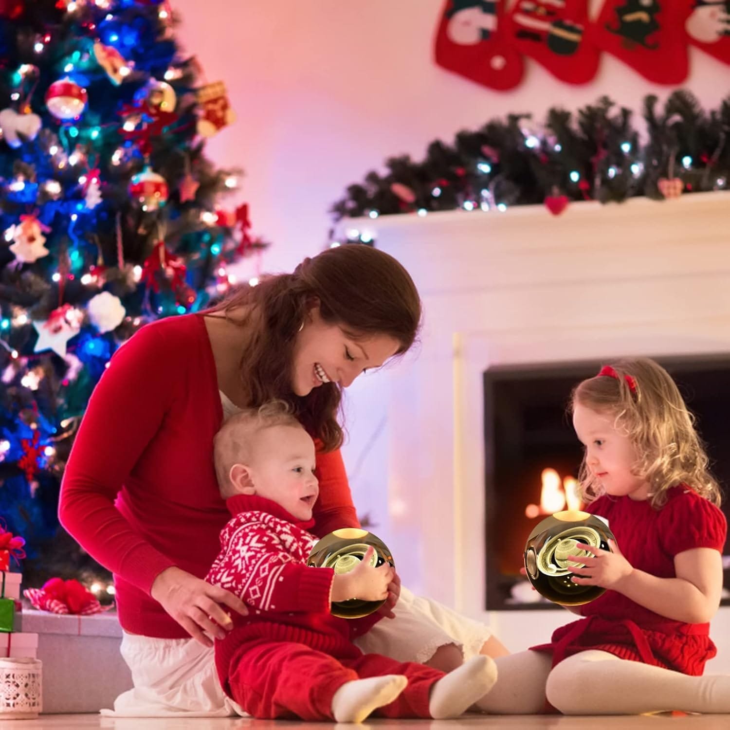 A woman and two children sit by a Christmas tree and fireplace, holding golden ornaments. They are dressed in red and white, surrounded by festive decorations.