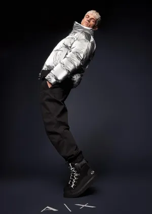 Person in a shiny silver puffer jacket and black pants leans backward at an angle against a dark background.