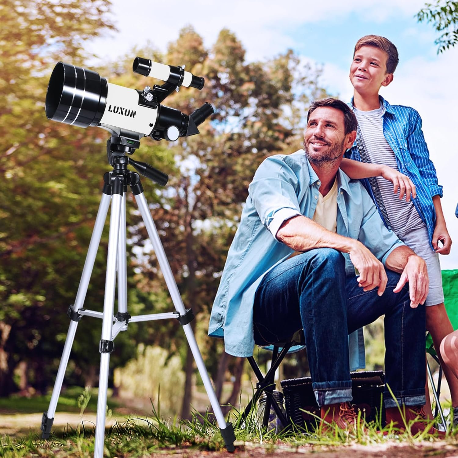 Man sitting and boy standing outdoors beside a telescope on a tripod. Trees and sky in the background.