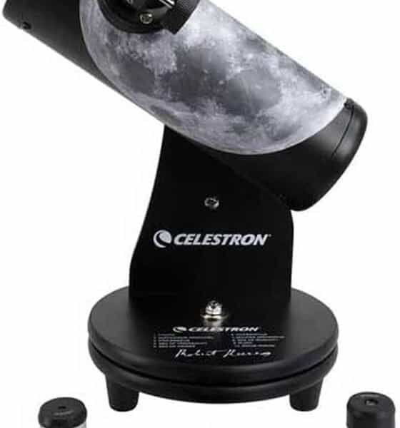 A tabletop Celestron telescope with a moon graphic on the tube, shown with two eyepieces and a lens cap on a white background.