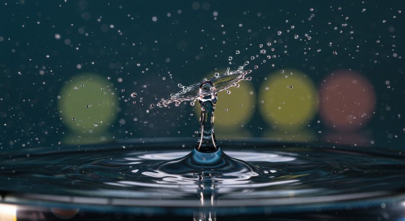 A close-up photo of a water droplet splashing and forming a crown shape above the water surface, with blurred colored lights in the background.