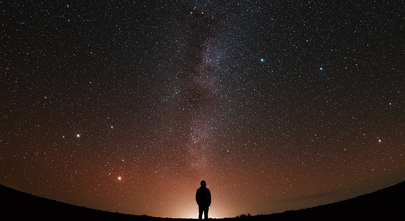 A person stands silhouetted against the horizon, looking up at a clear night sky filled with stars and the Milky Way.
