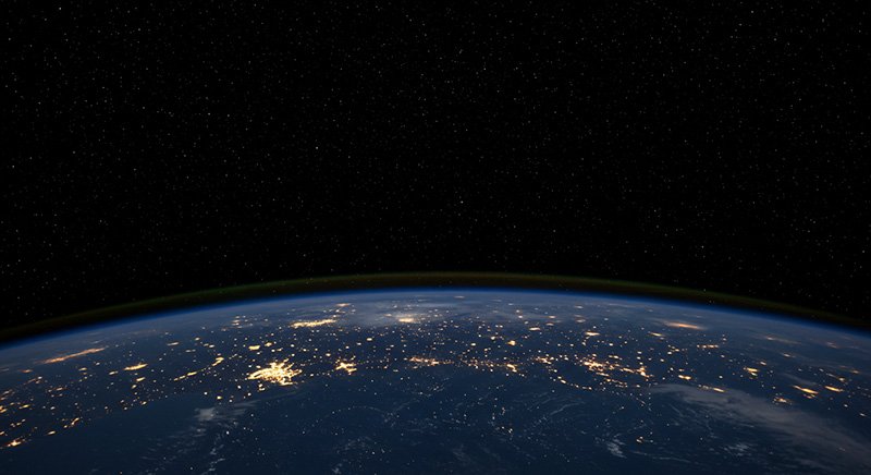 A view of Earth from space at night, showing illuminated cities and continents beneath a dark, star-filled sky.