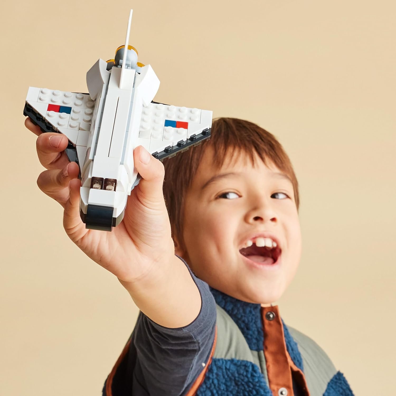A child holds up a toy spaceship made of interlocking plastic bricks, smiling with mouth open against a plain beige background.