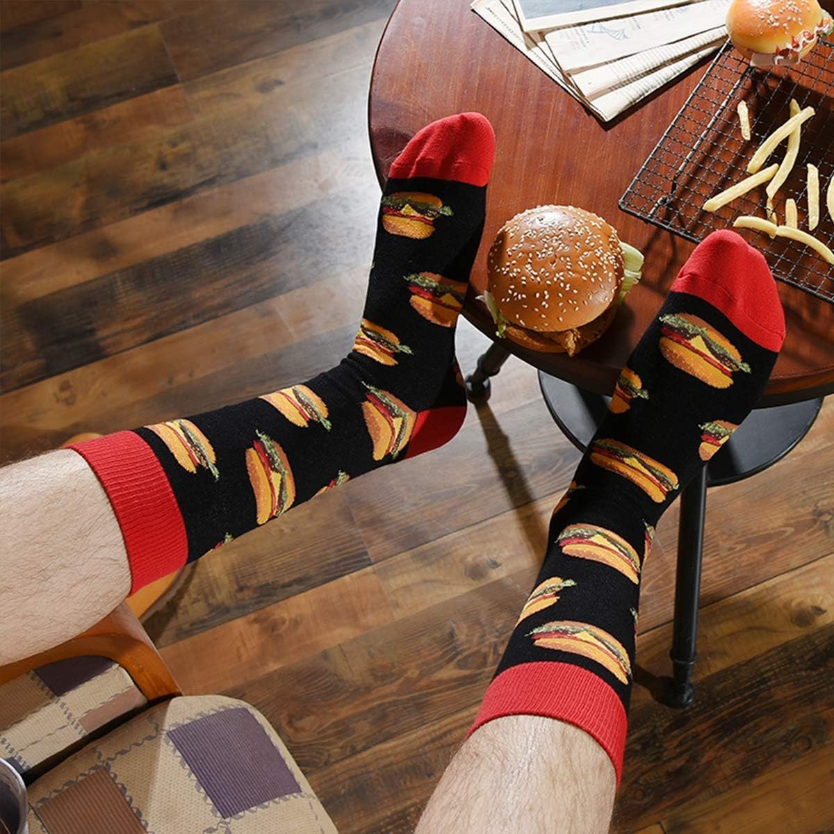 A person wearing black socks with hamburger patterns and red cuffs rests their feet near a table with a hamburger, fries, and newspapers.