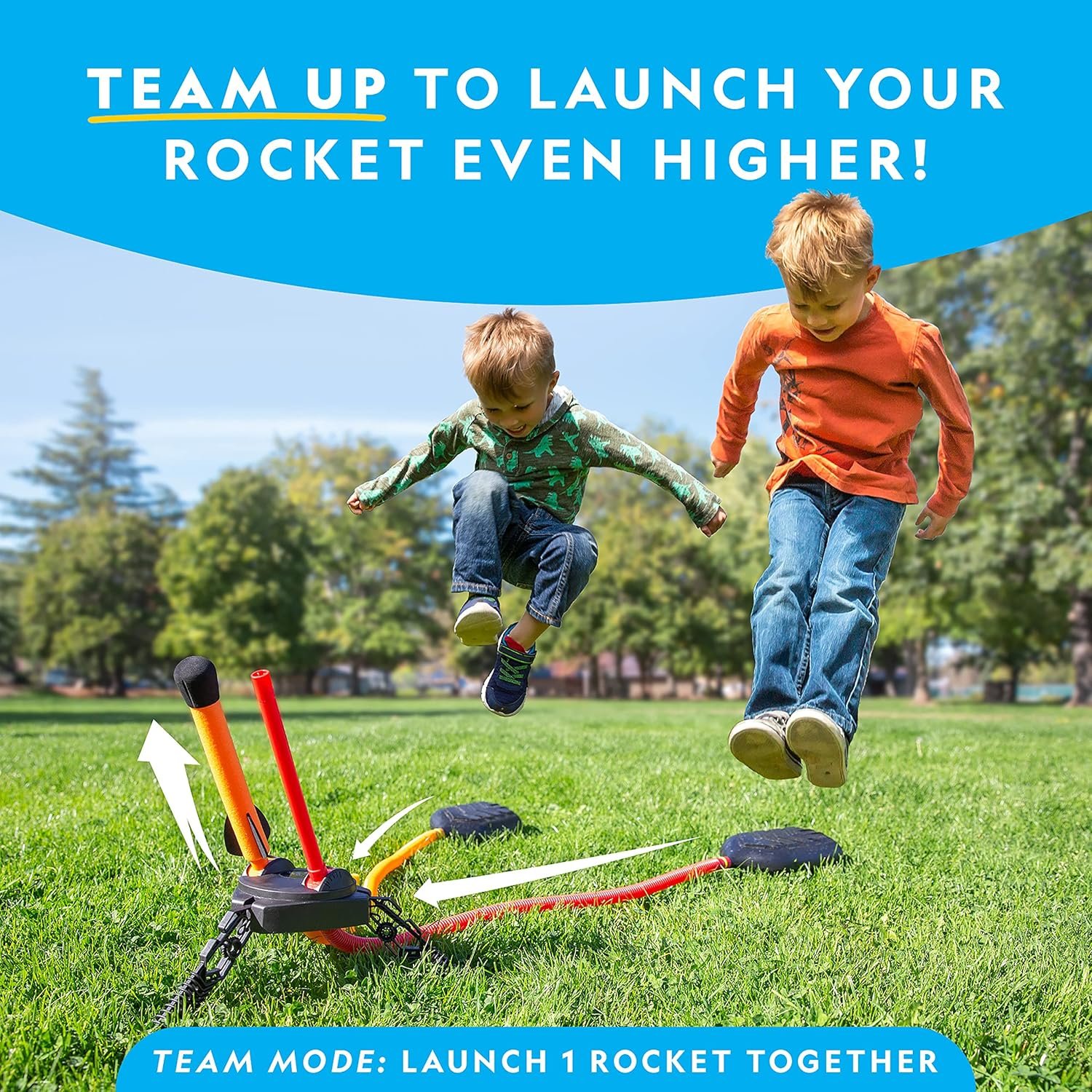 Two children jump onto a rocket launcher stomp pad in a grassy park, preparing to launch a toy rocket as part of a teamwork activity.