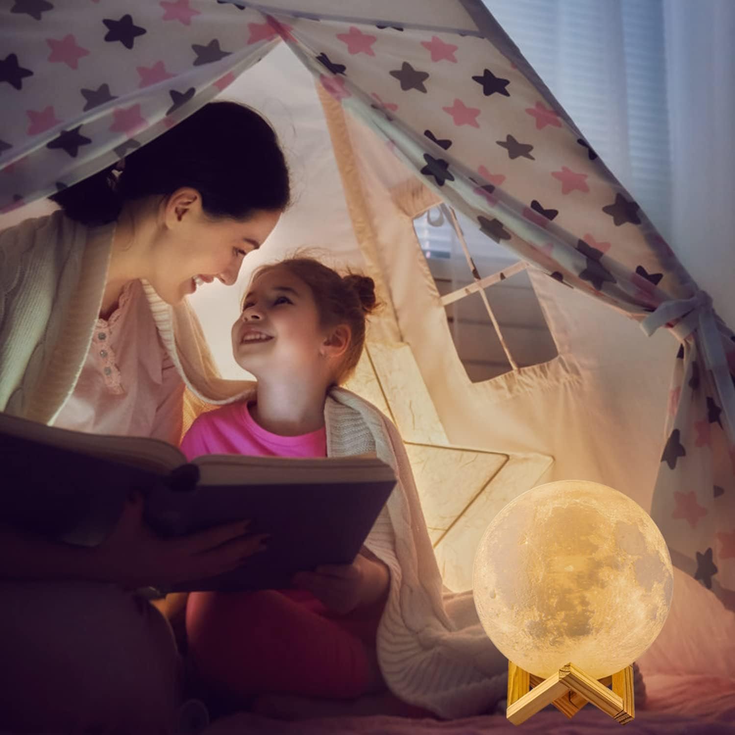 A woman and a girl sit inside a star-patterned tent, smiling at each other while reading a book next to a glowing moon-shaped lamp.