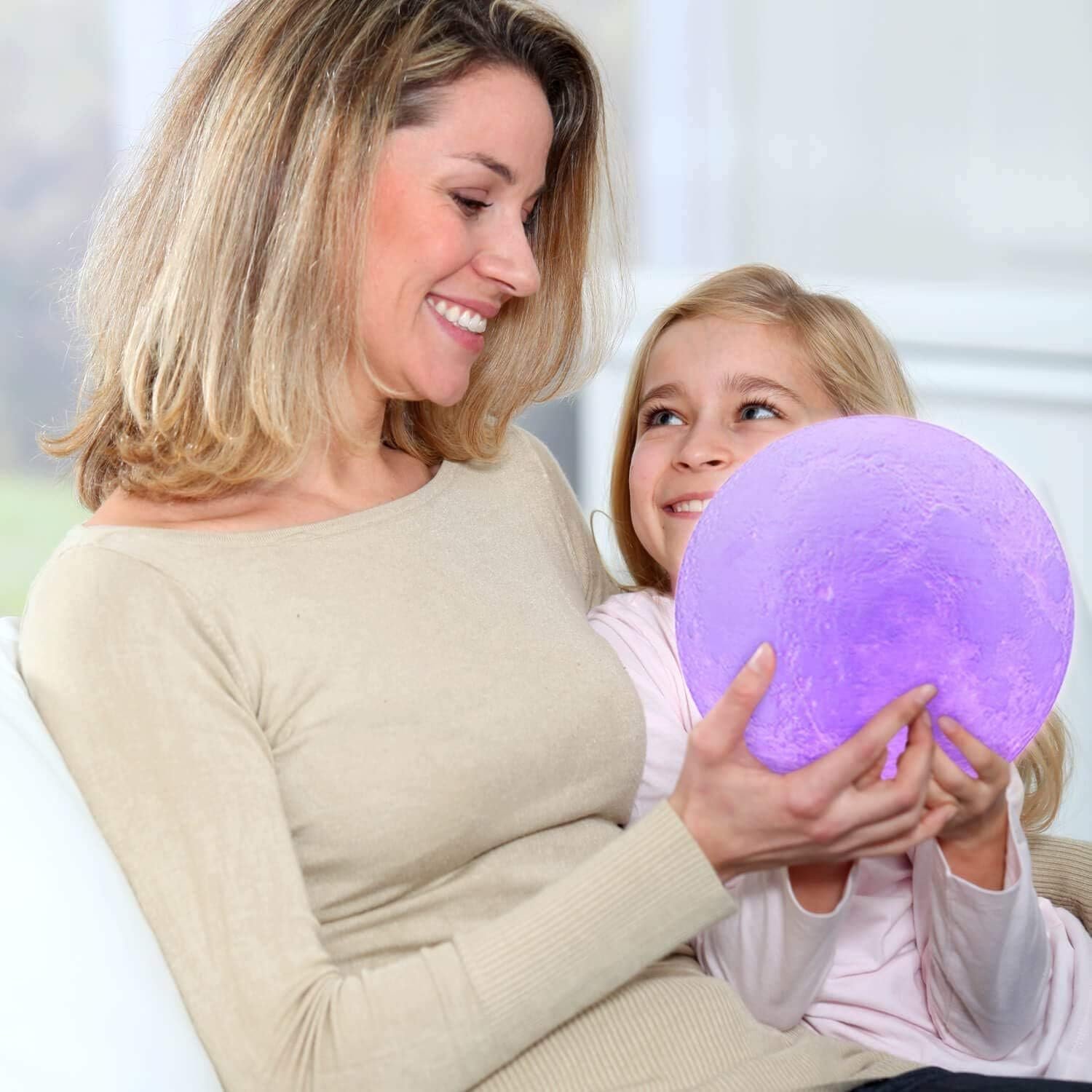 A woman and a young girl sit together on a sofa, smiling while holding a glowing purple sphere.
