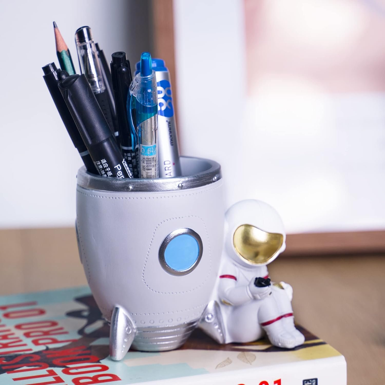A rocket-shaped pen holder with pens and markers inside, next to a small astronaut figurine, sits on top of a closed book on a wooden surface.