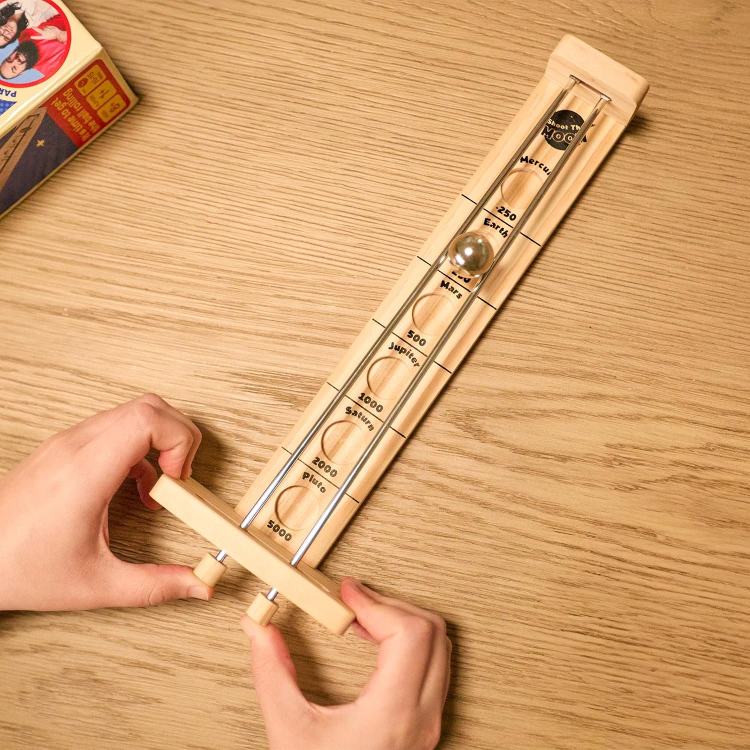 A person plays a wooden tabletop game, rolling a metal ball up a ramp marked with different point zones on a wooden surface.