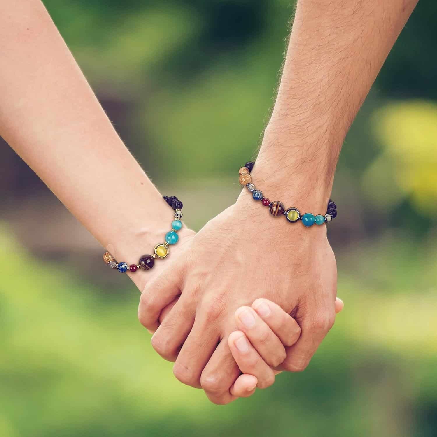 Two people holding hands, each wearing a beaded bracelet with multicolored stones, against a blurred green outdoor background.