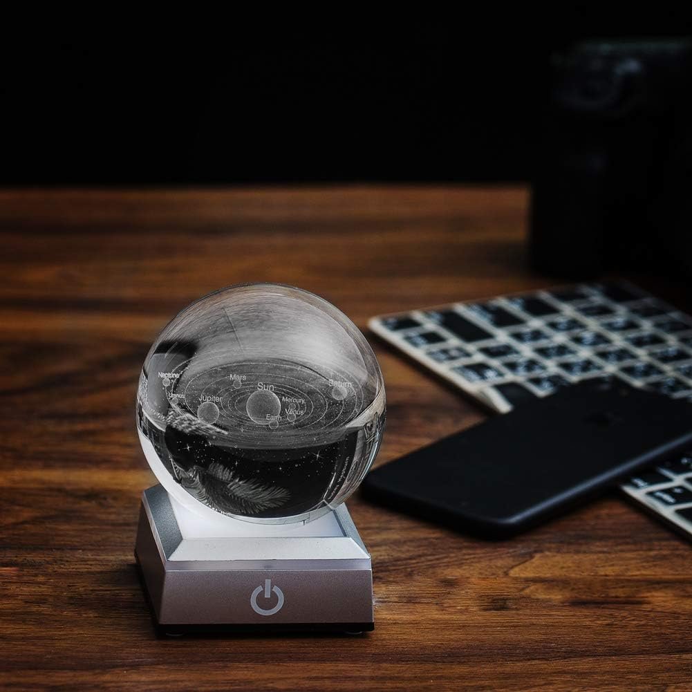 A glass sphere with an etched solar system model sits on a square lighted base, next to a black smartphone and a keyboard on a wooden surface.
