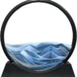 Round sand art sculpture with blue and white sand forming mountain-like patterns, encased in a circular black frame on a black base.