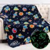 A navy blue throw blanket with colorful cartoon space-themed designs, including rockets, planets, and UFOs; a circular inset shows the blanket glowing with green outlines in the dark.