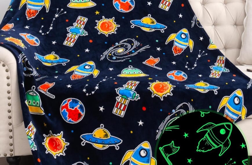 A navy blue throw blanket with colorful cartoon space-themed designs, including rockets, planets, and UFOs; a circular inset shows the blanket glowing with green outlines in the dark.