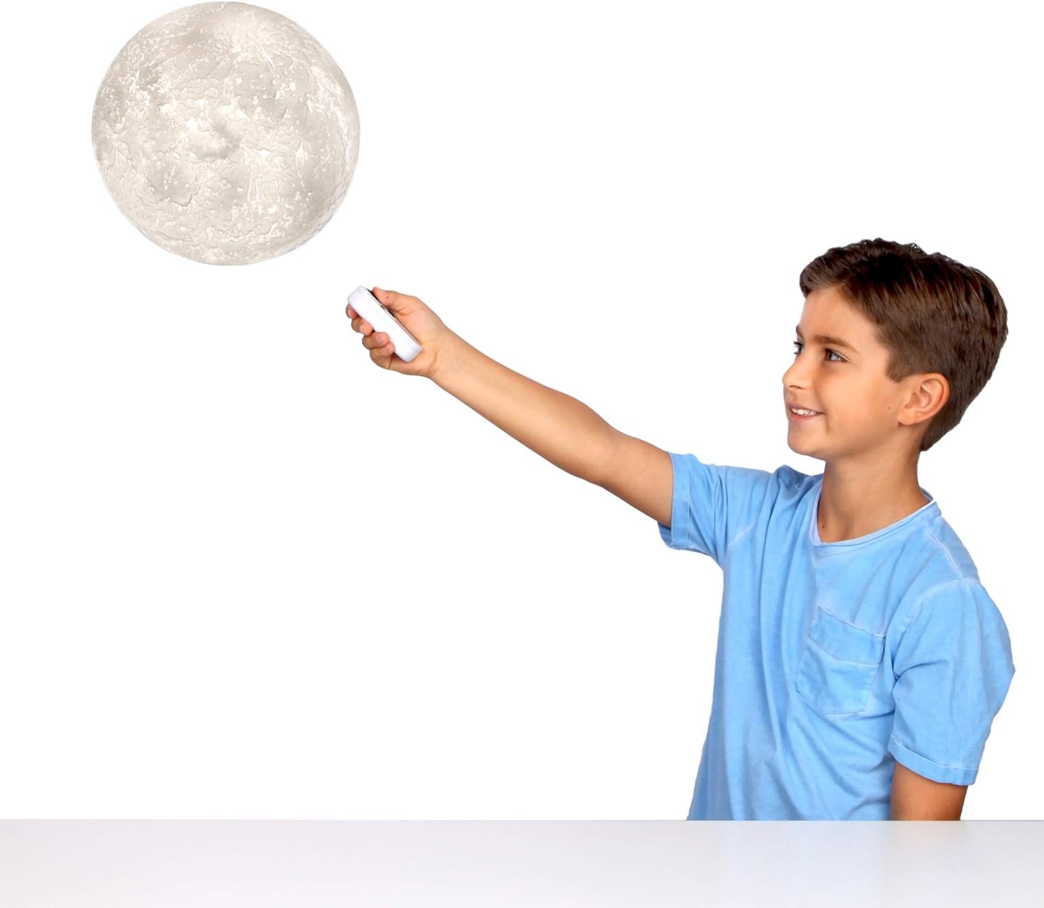 A boy in a blue shirt points a remote control at a large, illuminated moon model against a white background.