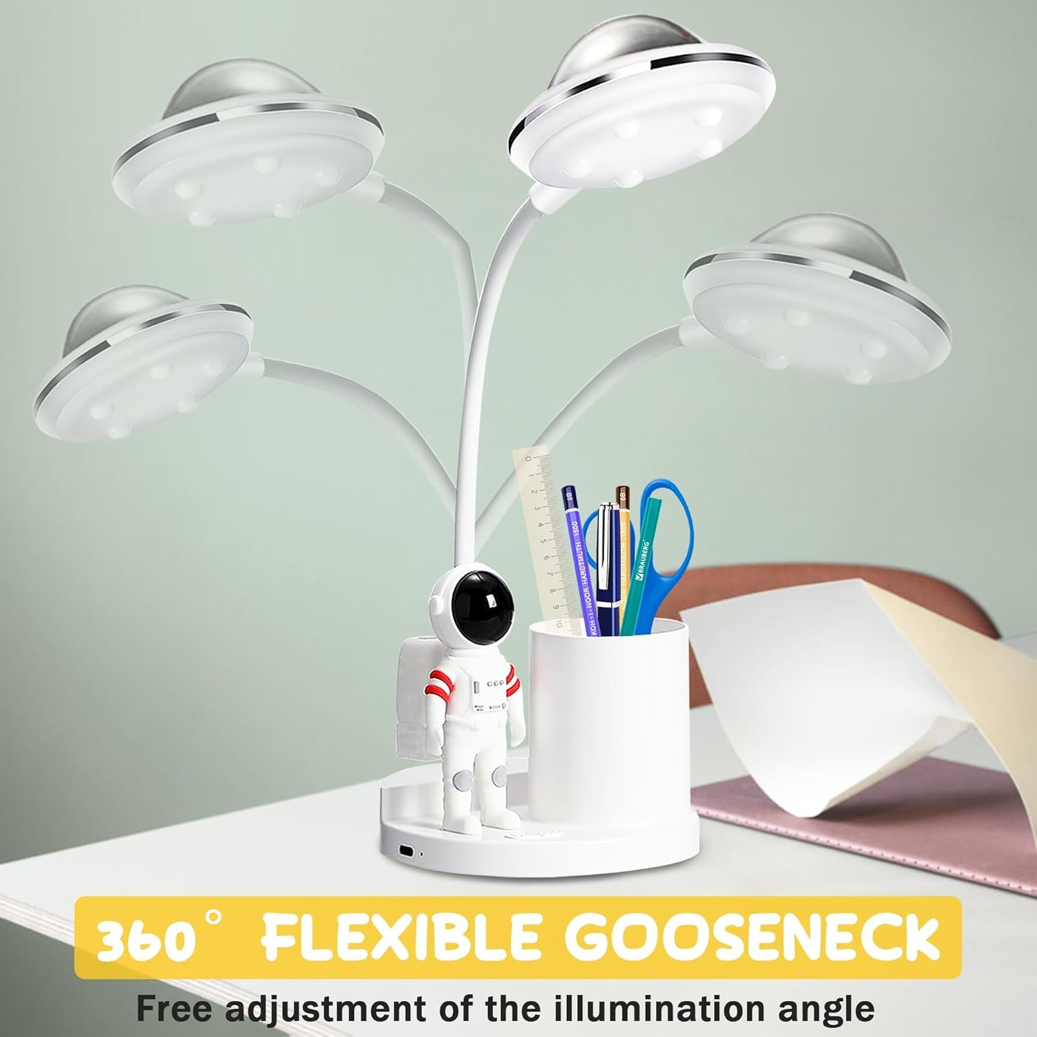 A white desk lamp with three UFO-shaped lights and a flexible gooseneck stands next to a pen holder designed like an astronaut, placed on a desk with scattered papers.