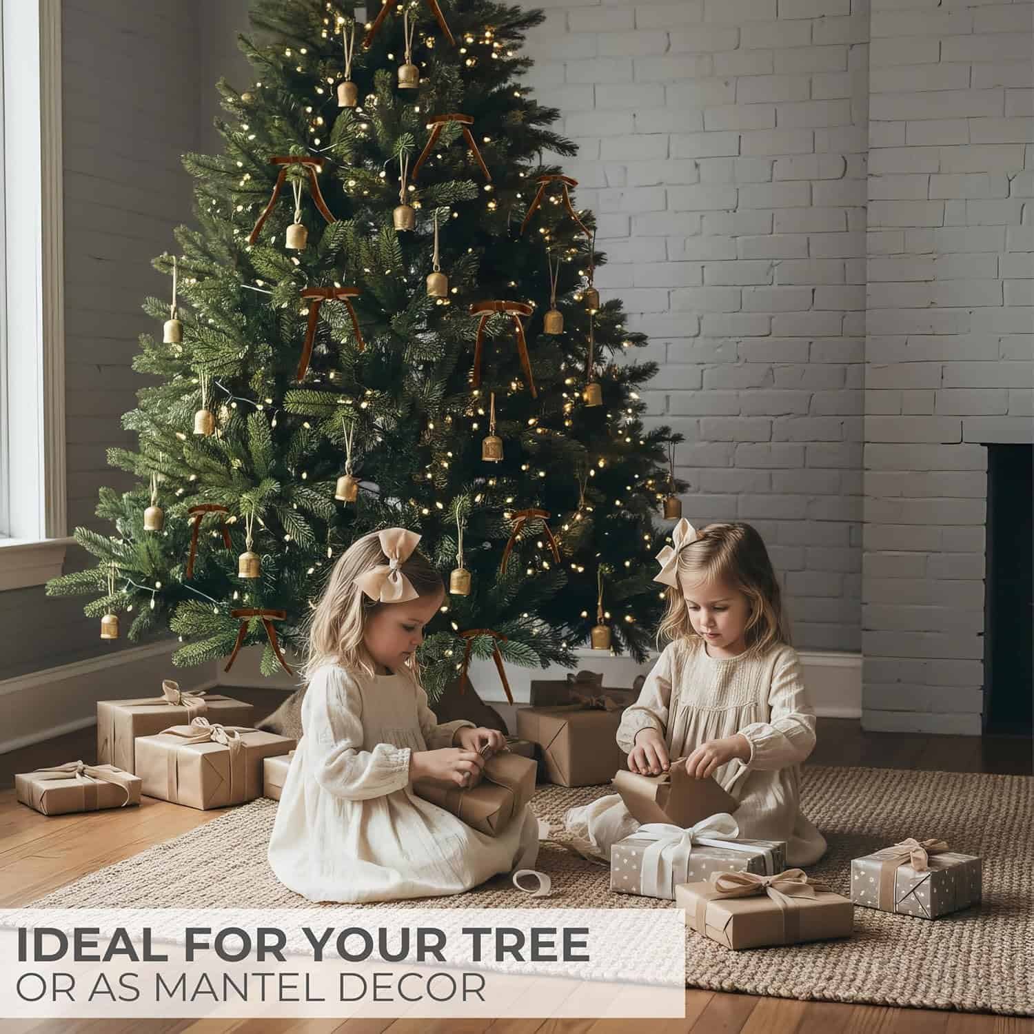 Two young girls in light-colored dresses sit on a woven rug, unwrapping presents in front of a decorated Christmas tree with string lights and gold ornaments.