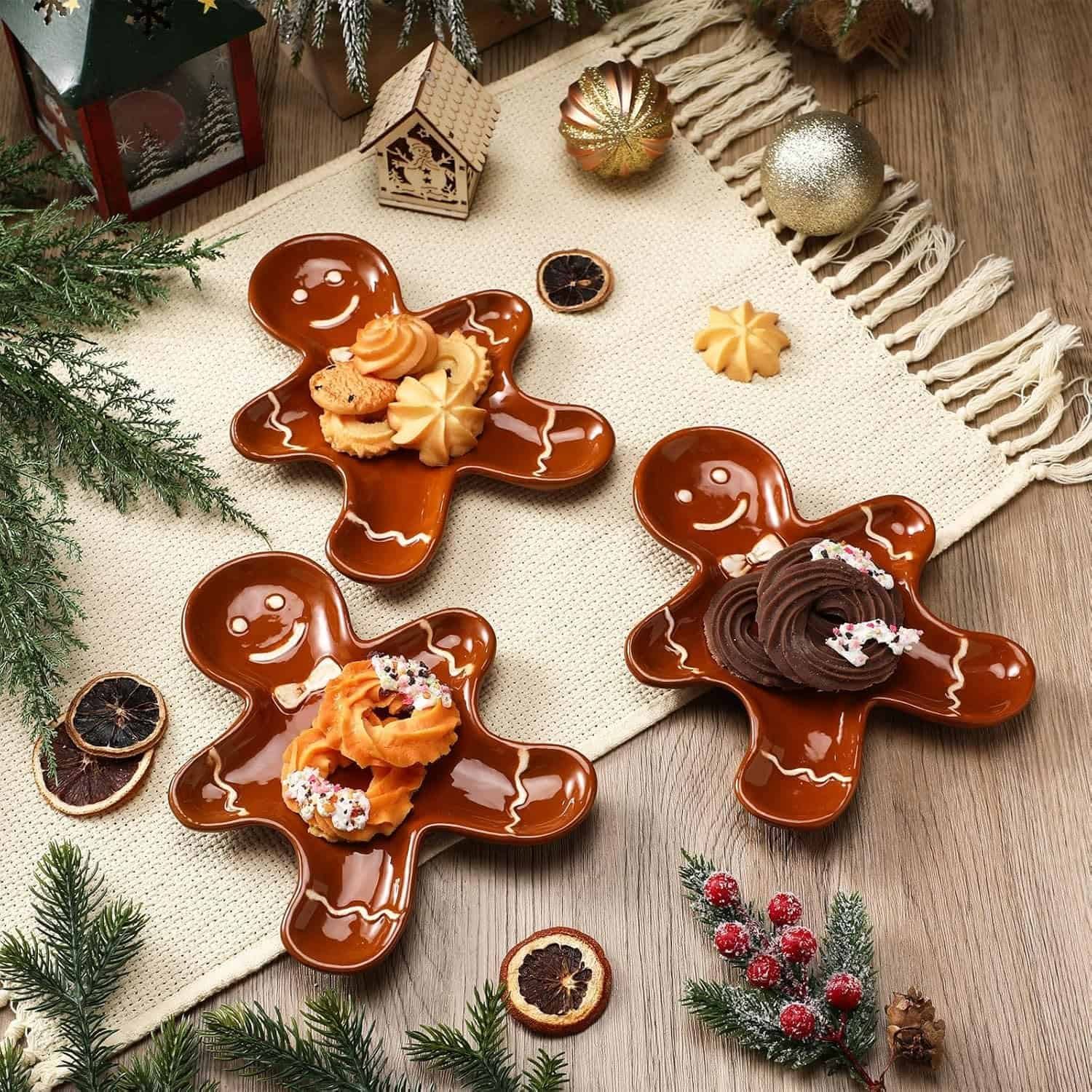 Three gingerbread man-shaped plates with assorted cookies are arranged on a beige placemat, surrounded by Christmas decorations and greenery on a wooden surface.