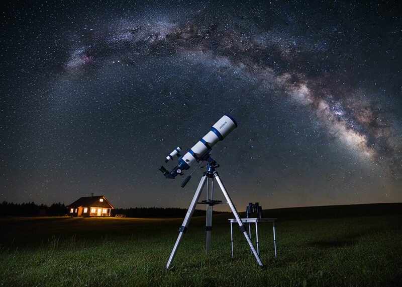 A large telescope on a tripod stands on grass at night, with a small lit house in the background and the Milky Way shining brightly in the star-filled sky above.