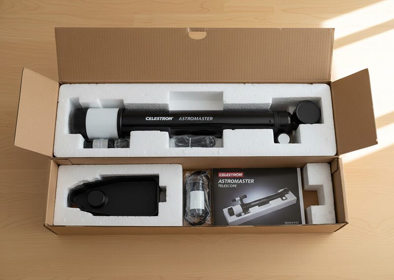 A Celestron AstroMaster telescope and accessories are neatly packed in a cardboard box with foam inserts, alongside an instruction manual for effortless telescope setup.