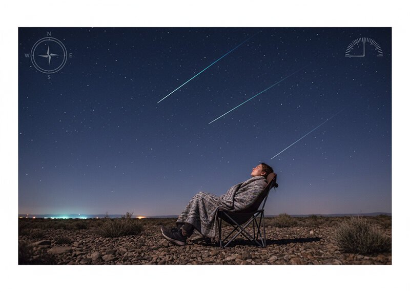 A person wrapped in a blanket sits on a chair outdoors at night, watching a spectacular meteor shower as multiple meteors streak across the starry sky.