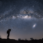 A silhouette of a person with a backpack looks at a star-filled night sky, marveling at the glowing galaxies overhead, while standing on uneven ground dotted with sparse bushes.