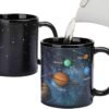 A hand pours hot water into a black mug, revealing a colorful solar system design with labeled planets. Another mug shows the plain black design with white stars.