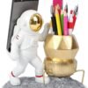 A desk organizer shaped like an astronaut and lunar lander, holding a smartphone and various pens, pencils, and a ruler on a gray, moon-like base.