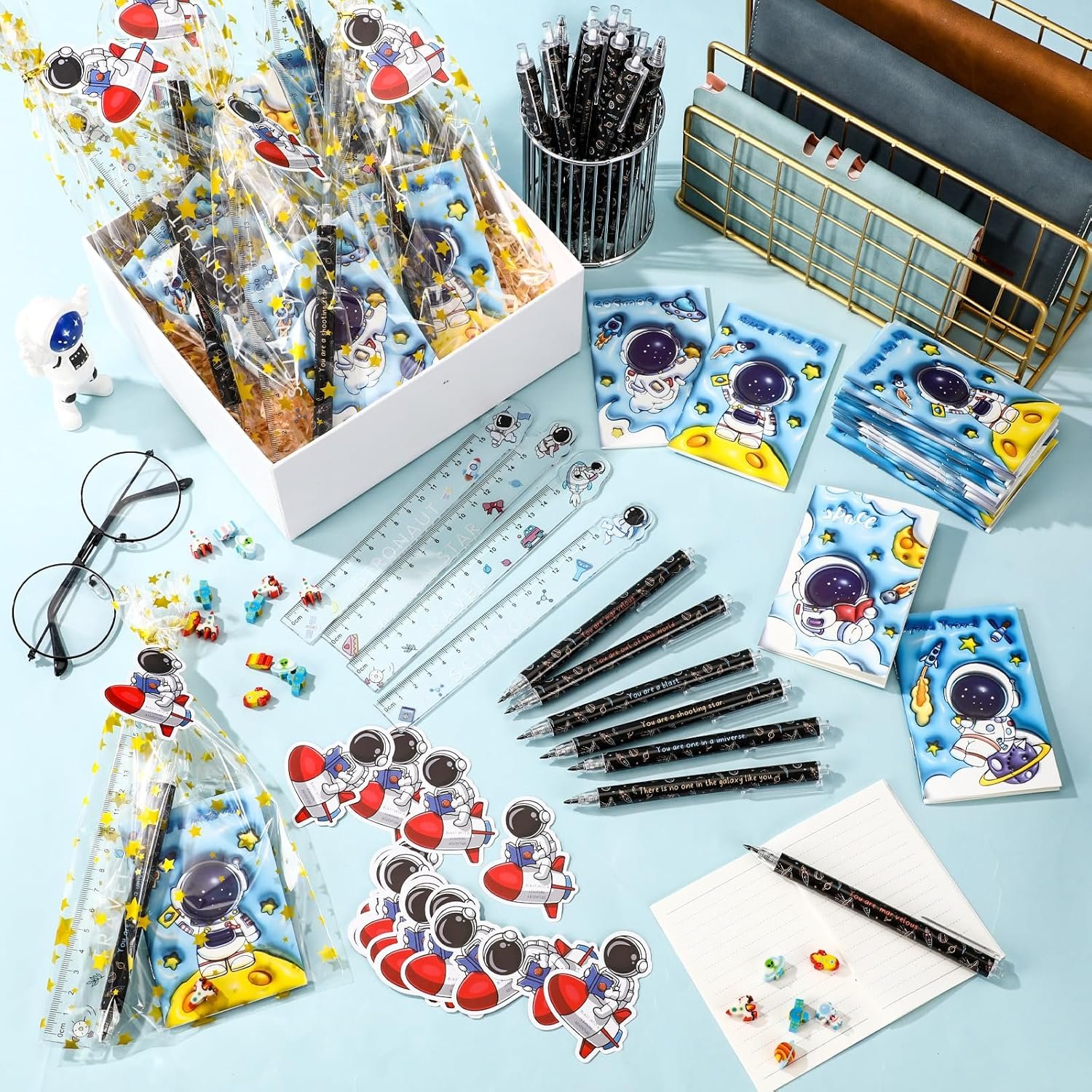 A collection of astronaut-themed stationery items, including pens, rulers, stickers, notepads, and gift bags, displayed on a light blue surface with a few small decorative items.