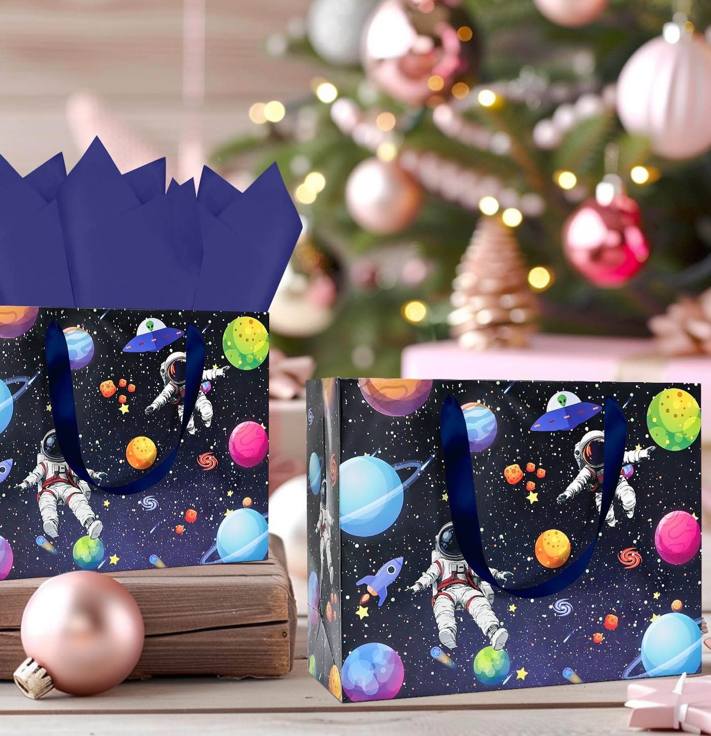 Two gift bags with space-themed designs featuring astronauts, planets, and UFOs are placed in front of a decorated Christmas tree with ornaments and wrapped gifts.