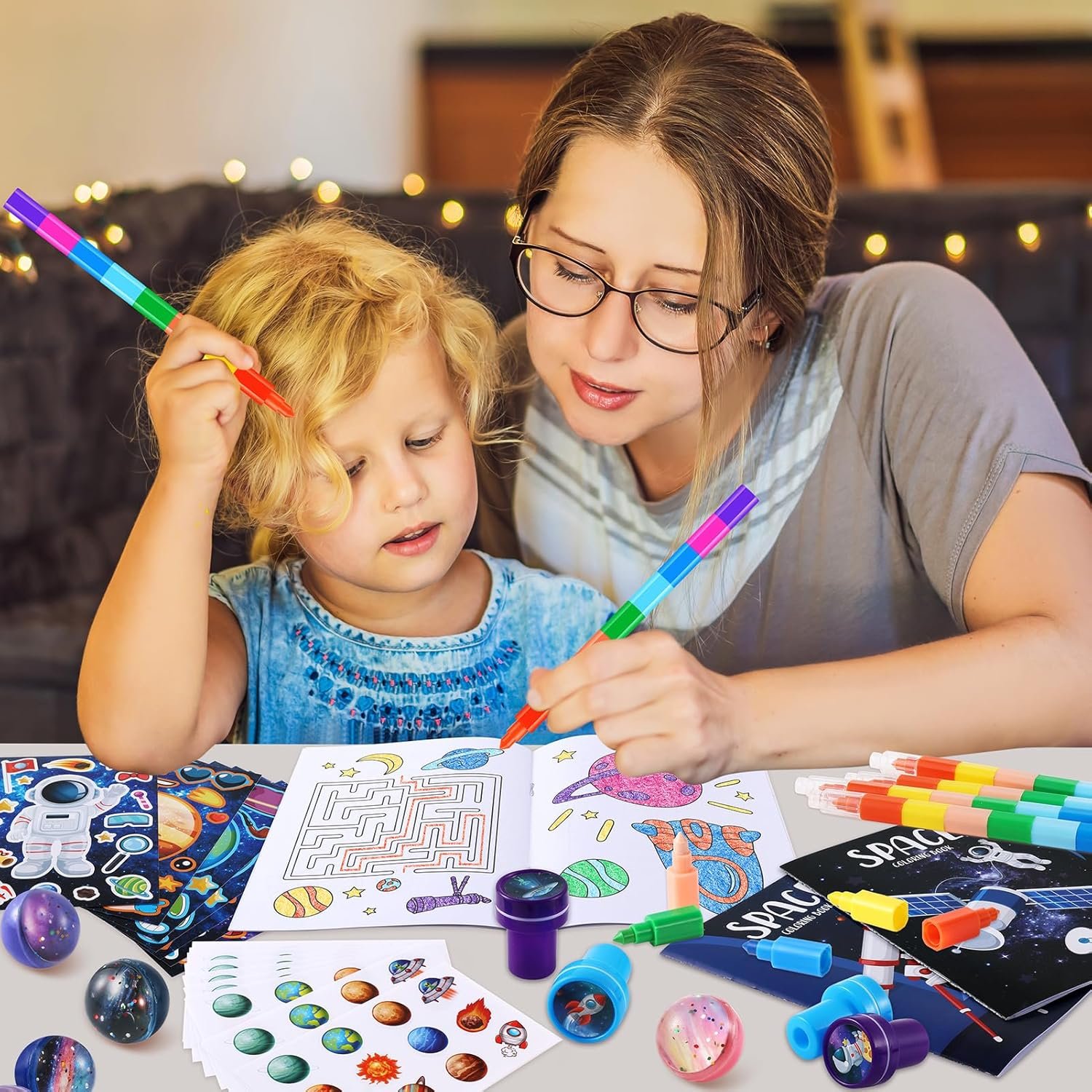 An adult and a child sit at a table coloring space-themed pictures with markers, surrounded by art supplies and space-themed stickers and toys.