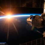 A NASA spacecraft docked to the International Space Station is seen with the sun rising over Earth's horizon in the background.