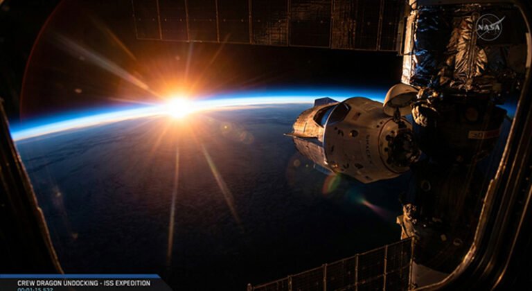 A NASA spacecraft docked to the International Space Station is seen with the sun rising over Earth's horizon in the background.